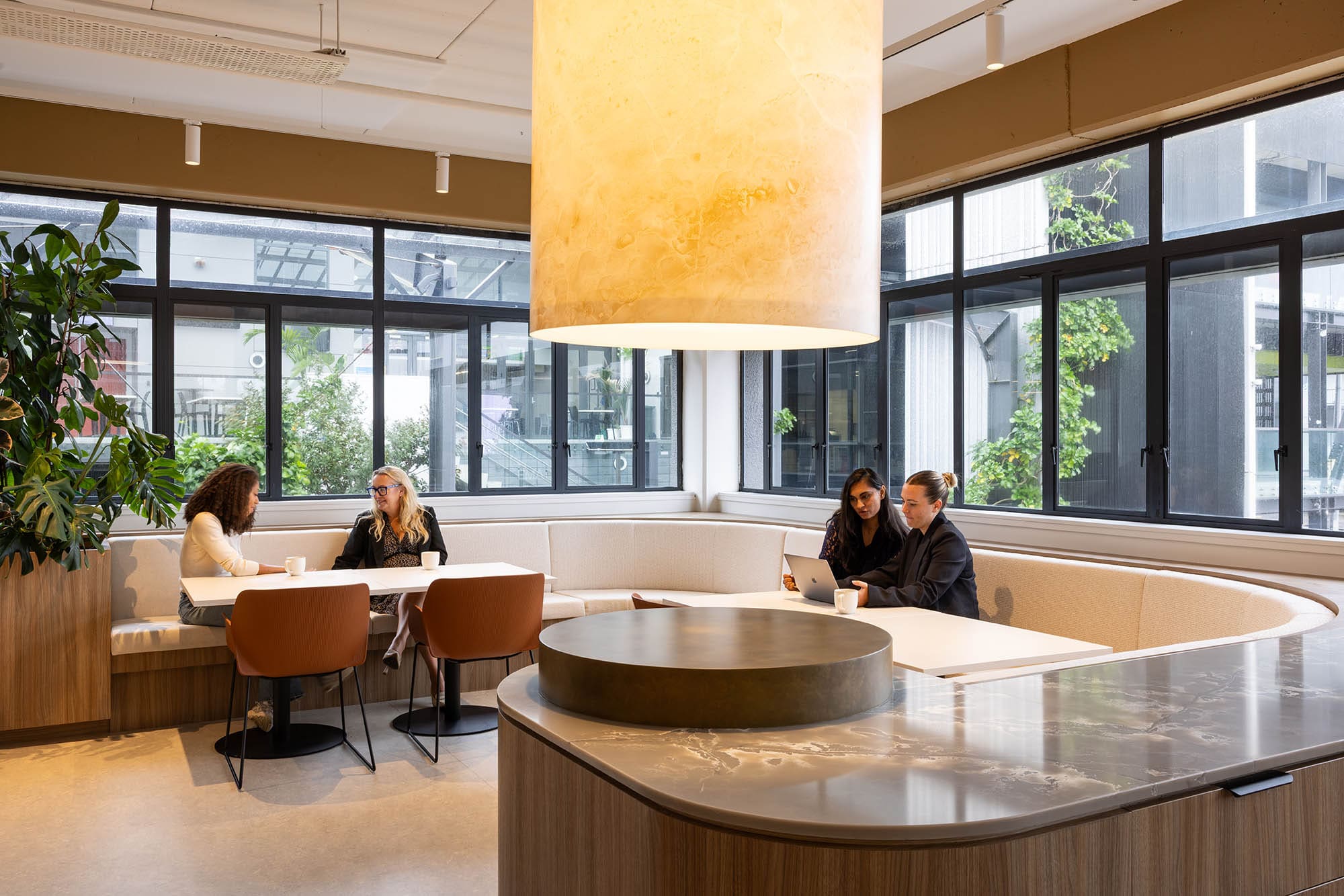 Two pairs of coworkers sit at booth tables in a bright, modern office lounge with large windows, a large hanging light fixture, and a central counter, talking and working on a laptop.