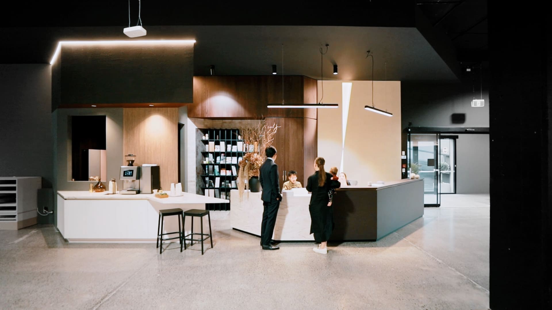 Modern, brightly lit retail interior featuring warm wooden wall panelling, minimalist open product shelving, and a clean concrete floor, with customers browsing and a staff member interacting at a central counter.